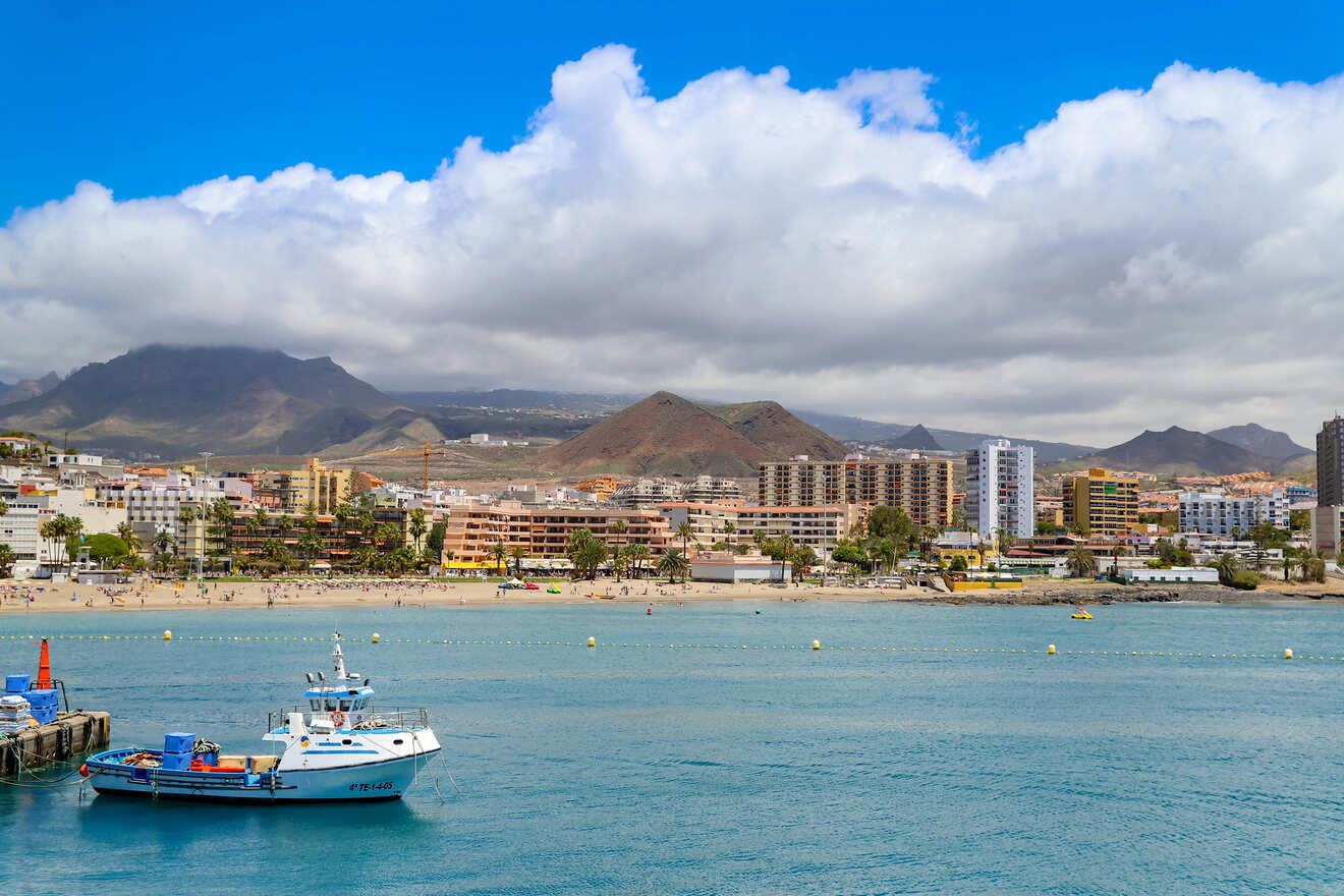 A panoramic view of a coastal cityscape with a sandy beach and a fishing boat docked in the harbor, with mountains under a partly cloudy sky