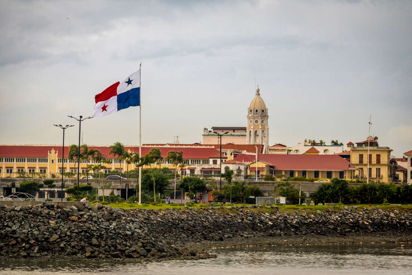 Scenic view of the Panamanian flag waving in front of the historic yellow and white Palacio de las Garzas building under a cloudy sky.