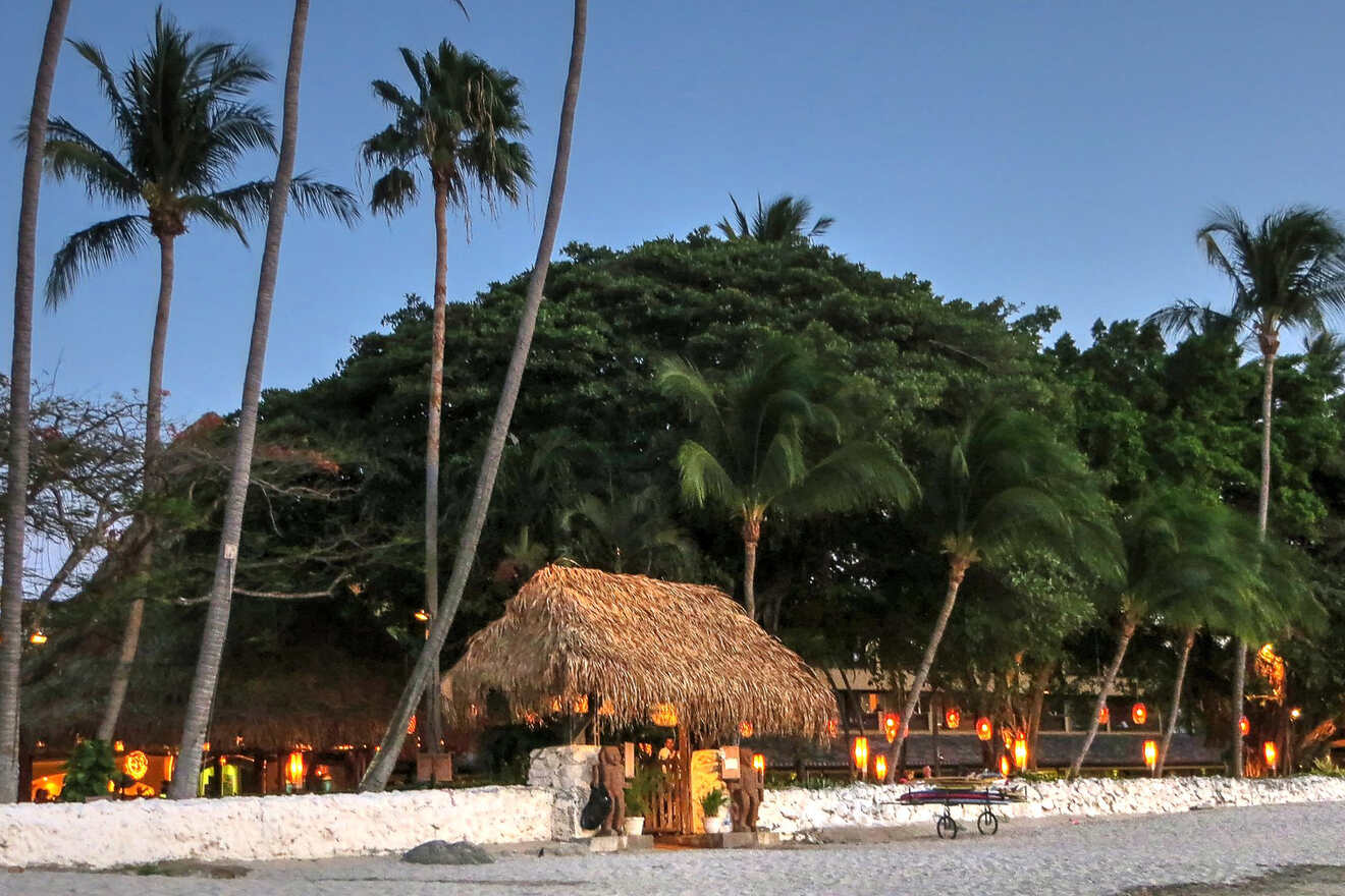 A tropical beachfront scene at dusk with palm trees, a thatched hut, and lantern-lit buildings nestled among lush greenery. A small cart is visible on the sand in the foreground.