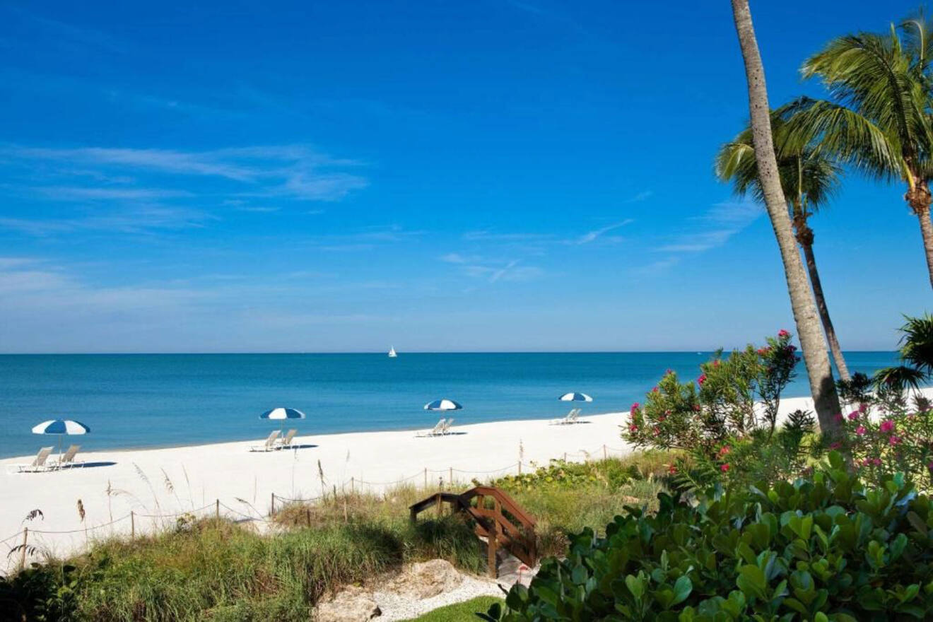 Scenic beach view with white sand and calm turquoise waters, framed by tropical foliage and palm trees, with thatched sun umbrellas and lounging chairs inviting relaxation