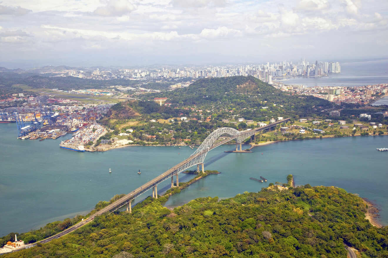 Aerial view of the Bridge of the Americas in Panama, spanning the entrance to the Panama Canal, with the city's skyline in the distance and lush greenery surrounding the area