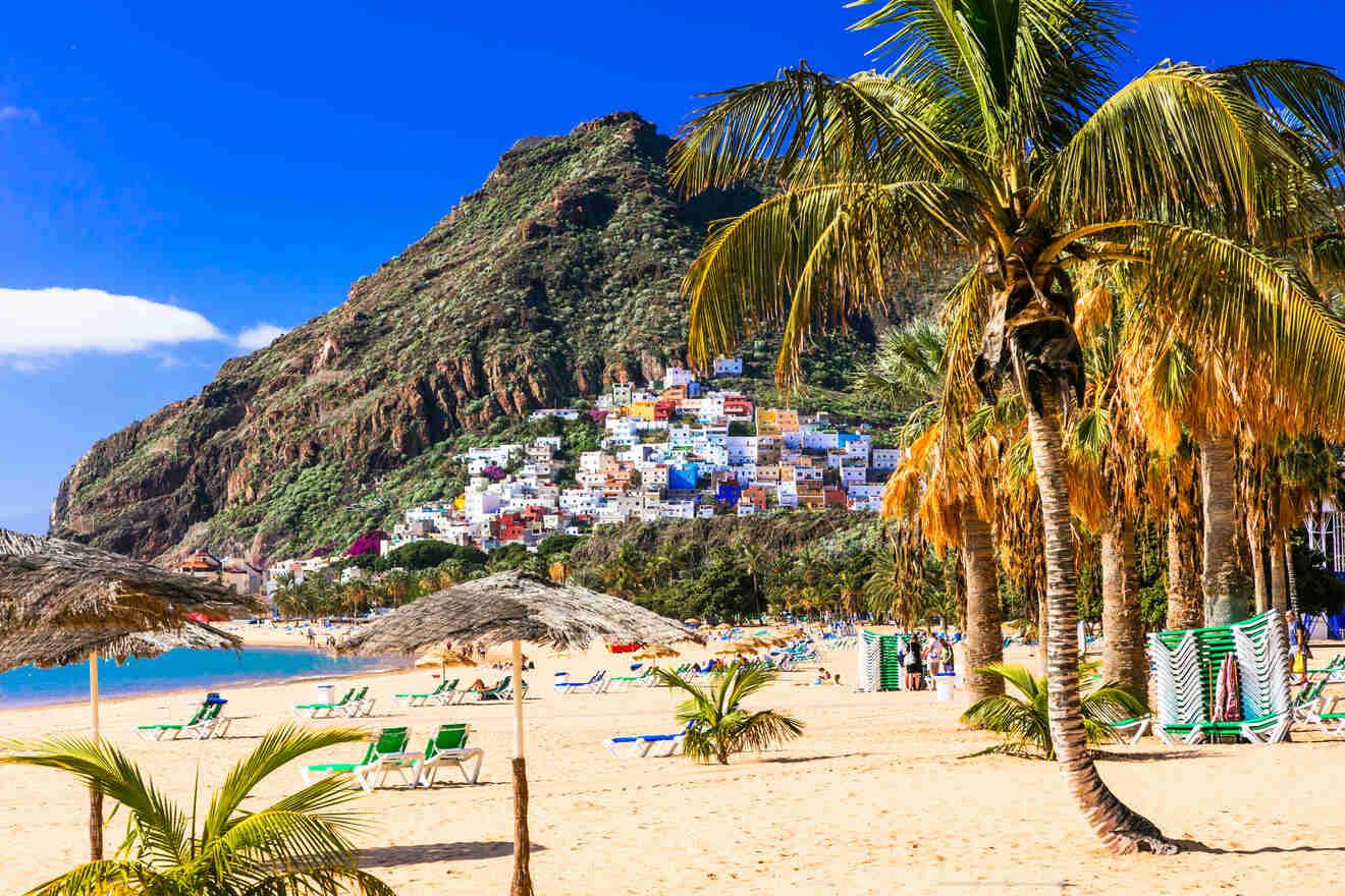 Scenic view of a colorful hillside village overlooking a sandy beach with palm trees and beachgoers enjoying the sunny day.