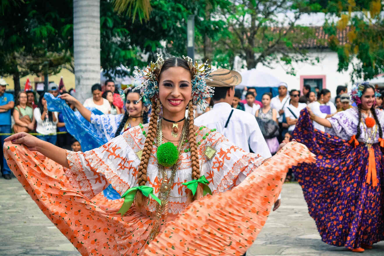 A woman in traditional bright floral attire and headpieces dances outdoors, accompanied by others in similar clothing. A crowd watches in the background. Trees and buildings are visible.