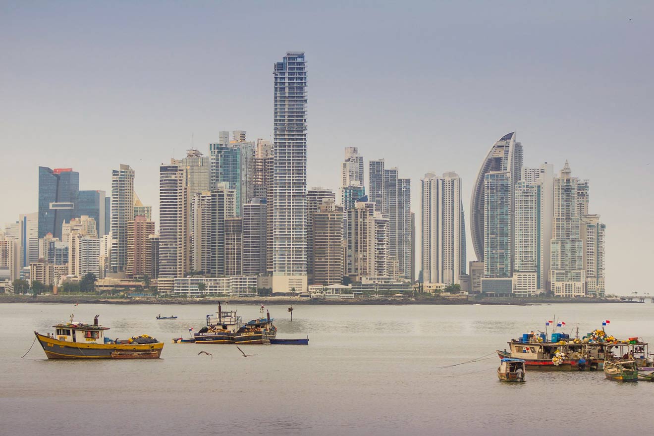 anoramic view of Panama City's skyline from across the water, with high-rise buildings reflecting the sunlight and fishing boats moored in the foreground