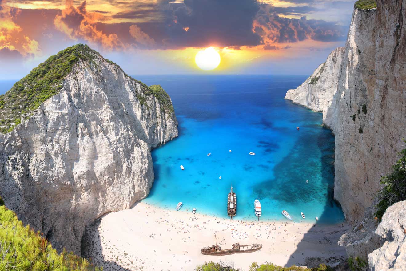 Aerial view of a beach cove with a shipwreck, clear blue water, boats, sandy shores, and surrounding cliffs at sunset.