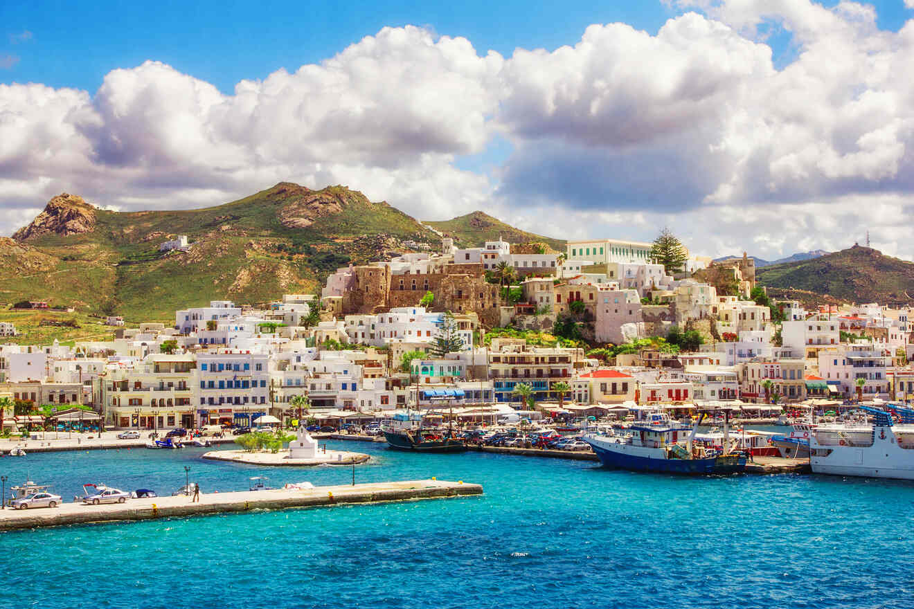 A picturesque coastal town with white buildings and colorful accents nestled against green hills and a bright blue sea under a partly cloudy sky.