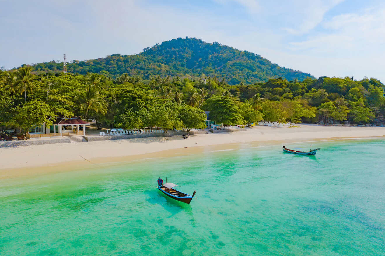 Aerial view of a tranquil beach in Thailand with a single traditional boat floating on clear turquoise water, lush greenery in the background, and a hint of beachfront resorts.

