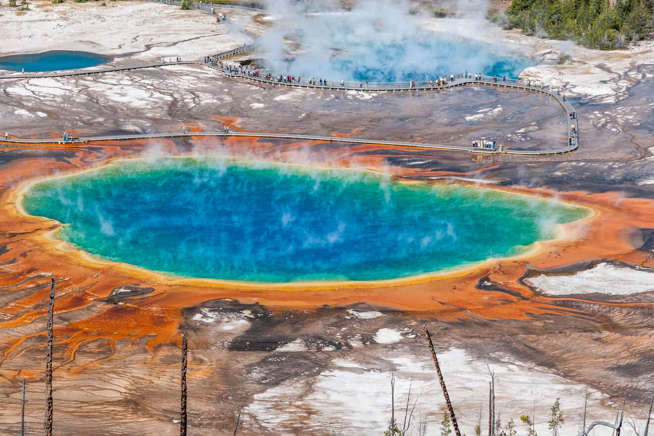 Aerial view of a large, colorful hot spring with vibrant blue, green, and orange hues, surrounded by boardwalks and visitors at Yellowstone National Park.