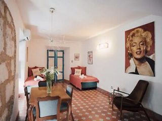 A neatly arranged room with two single beds, a dining table with chairs, a lounge chair, and a large painting of a woman on the wall. The floor has a red and white patterned design.