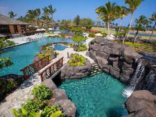 A resort pool with rock formations, a waterfall, and surrounding palm trees, featuring a wooden bridge and lounge chairs by the water.