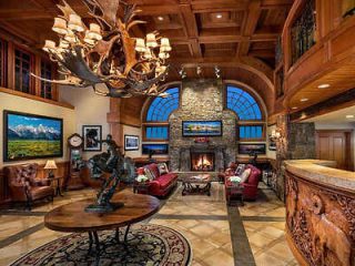 Luxurious living room with wooden ceiling, stone fireplace, leather couches, antler chandelier, and artwork on walls. A round table and decorative sculptures are in the center.