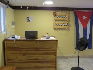 A small office space with a wooden desk, a laptop, a phone, a Cuban flag on the wall, and shelves holding small items. A fan stands on the floor. A small picture is also hanging on the wall.