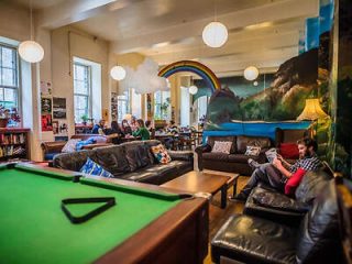 A  lively hostel common room painted with murals and a rainbow, furnished with black leather sofas and a pool table