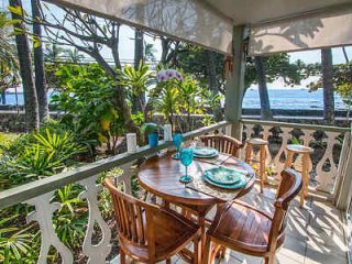 Outdoor dining area with a wooden table and chairs on a porch overlooking the ocean. Surrounded by lush greenery, with table set for four.