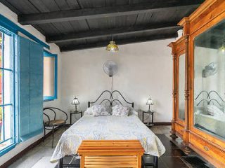 A bedroom features a metal bed with patterned bedding, a wooden wardrobe, a bench at the foot of the bed, two bedside tables with lamps, and a window with blue shutters.