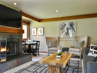 Cozy living room with a fireplace, wooden coffee table, two armchairs, a sofa, and a dining table. Wall art and a large TV are mounted above the fireplace.