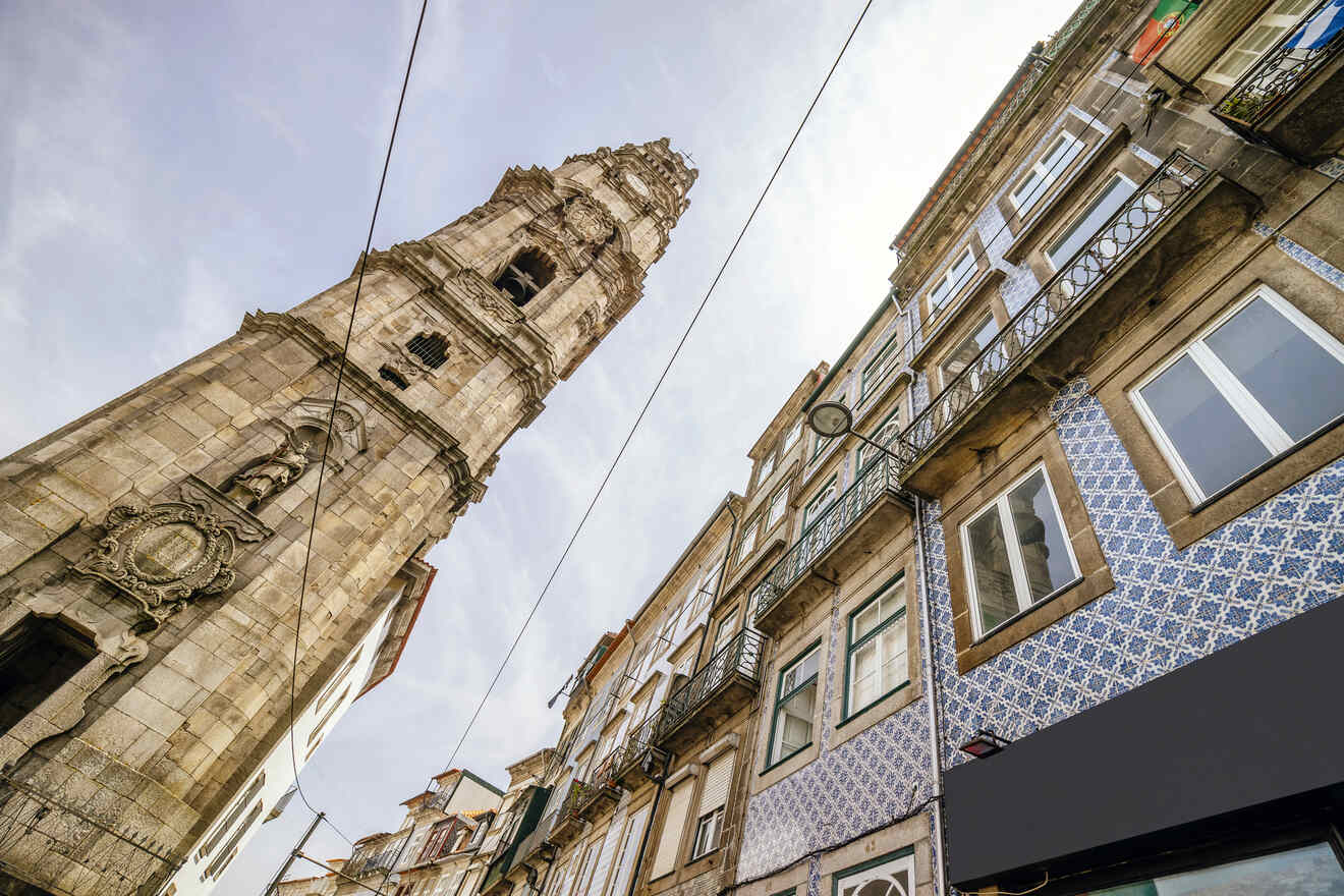 Looking up at the ornate Clérigos Tower in Porto, Portugal, with traditional Portuguese tiled facades of surrounding buildings framing the sky