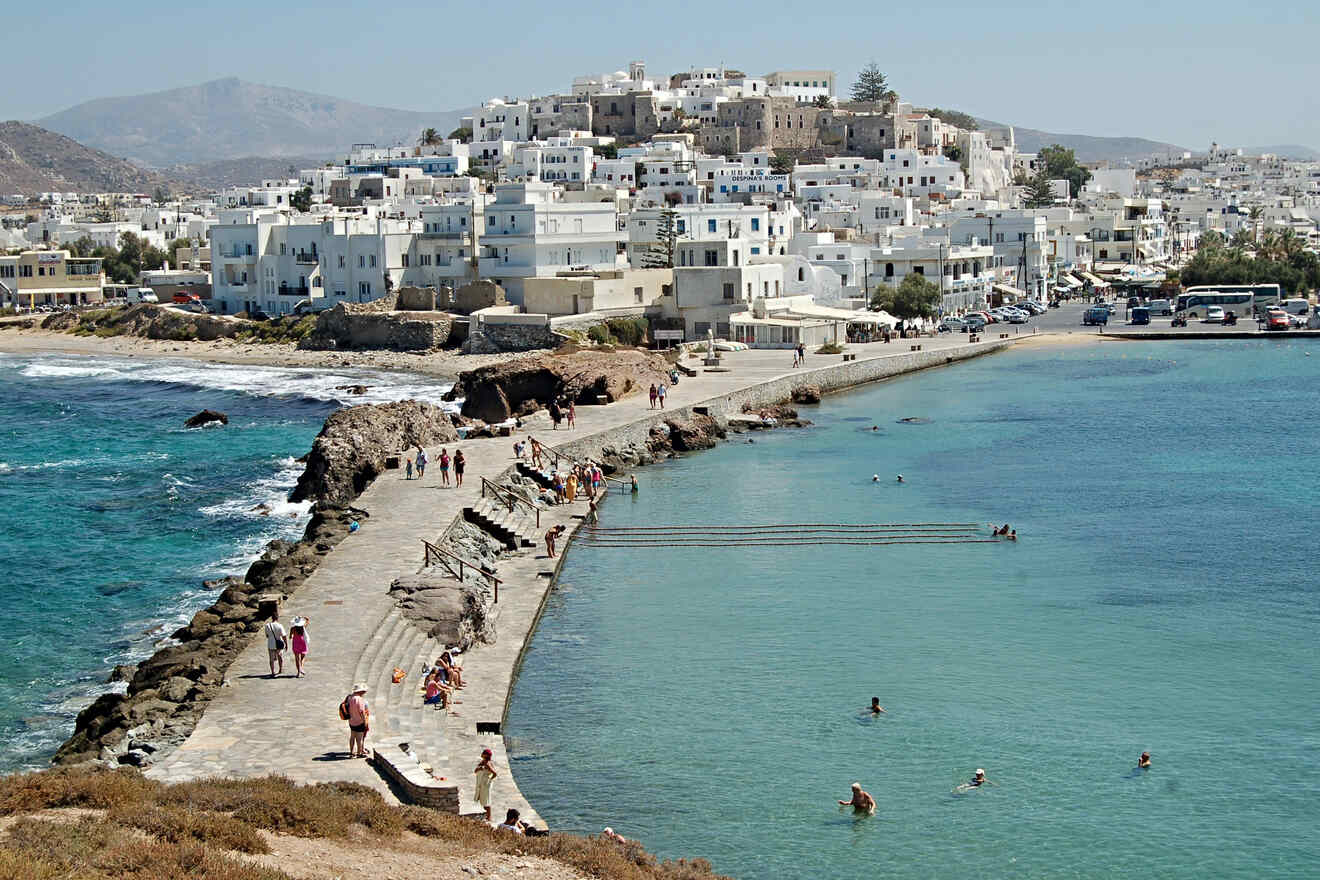 A scenic coastal walkway with people strolling and swimming, leading to a town with white buildings against a mountainous backdrop.