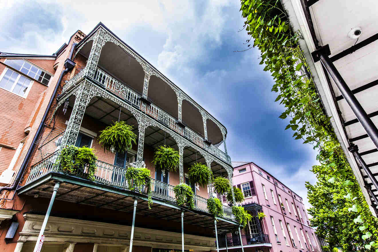 A three-story building with ornate wrought-iron balconies adorned with hanging ferns, set against a cloudy sky. Another building and greenery are visible on either side.