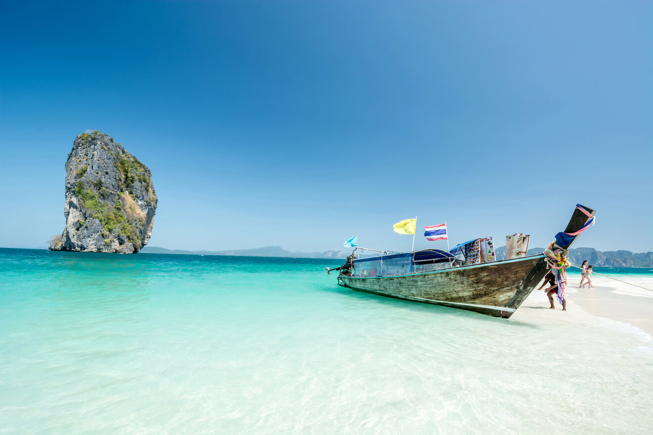 Pristine beach scene featuring a traditional long-tail boat on the shore of a clear turquoise sea with a magnificent limestone karst in the background, exemplifying Thailand's natural coastal beauty.

