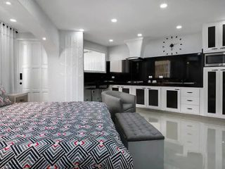 Modern studio apartment with a kitchen, bed, and lounge area. The room features black and white decor, geometric bedspread, flat-screen TV, and reflective floor tiles. A large wall clock is visible.