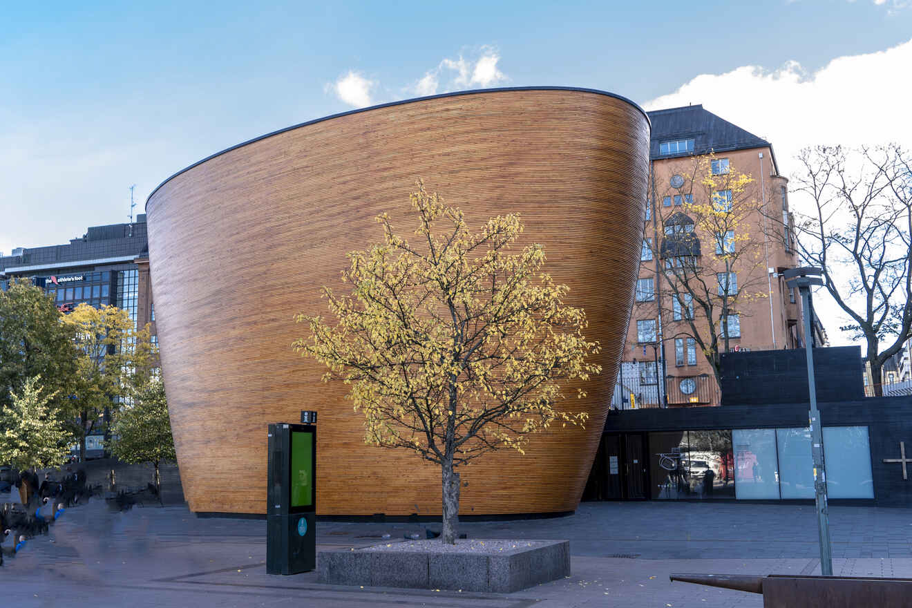 The curved wooden facade of the Kamppi Chapel of Silence in Helsinki stands out amidst a cityscape, juxtaposed with an autumnal tree in the foreground