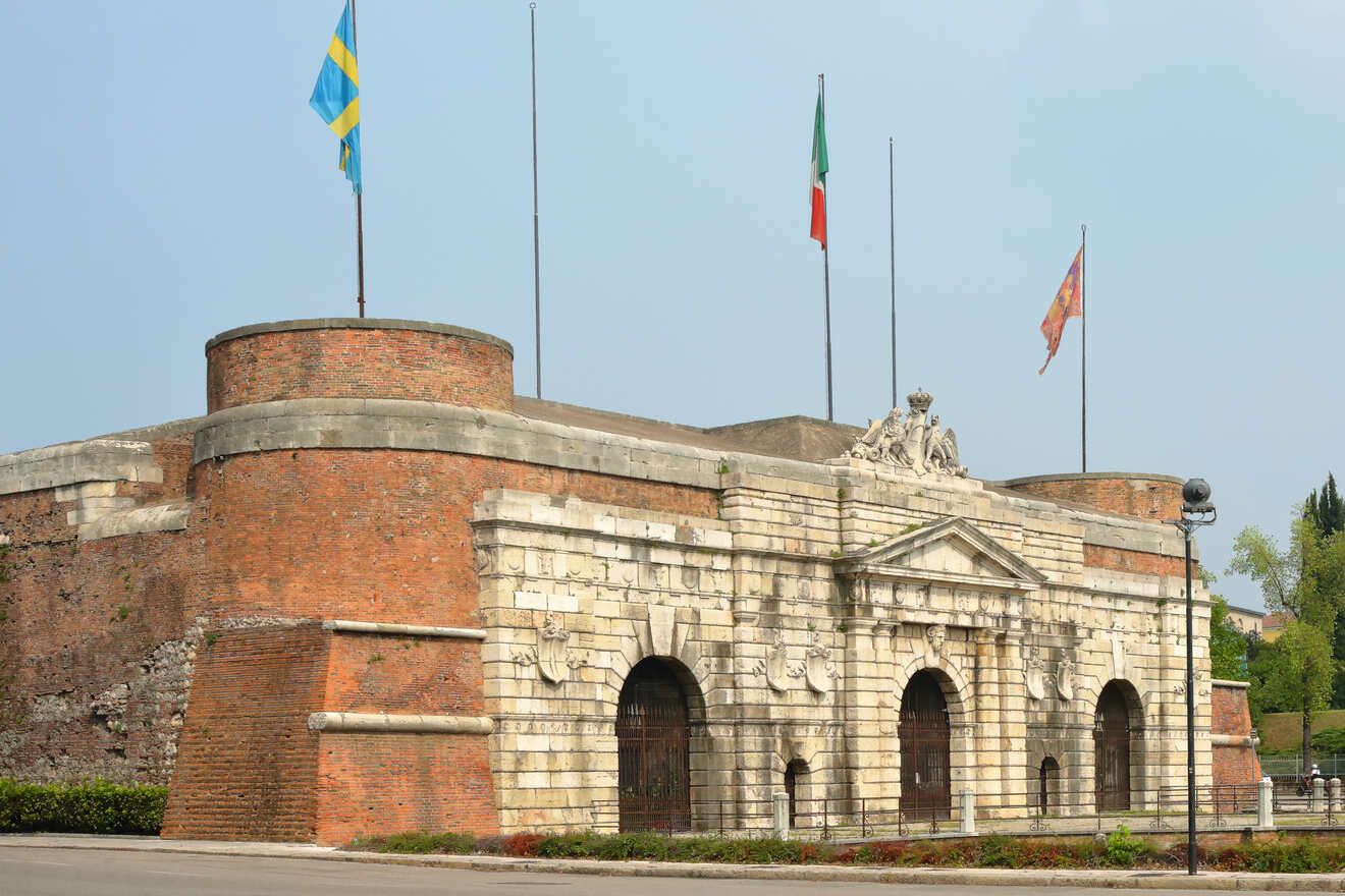 An ancient fortress entrance with brick walls, adorned with sculptures and national flags, under a clear sky