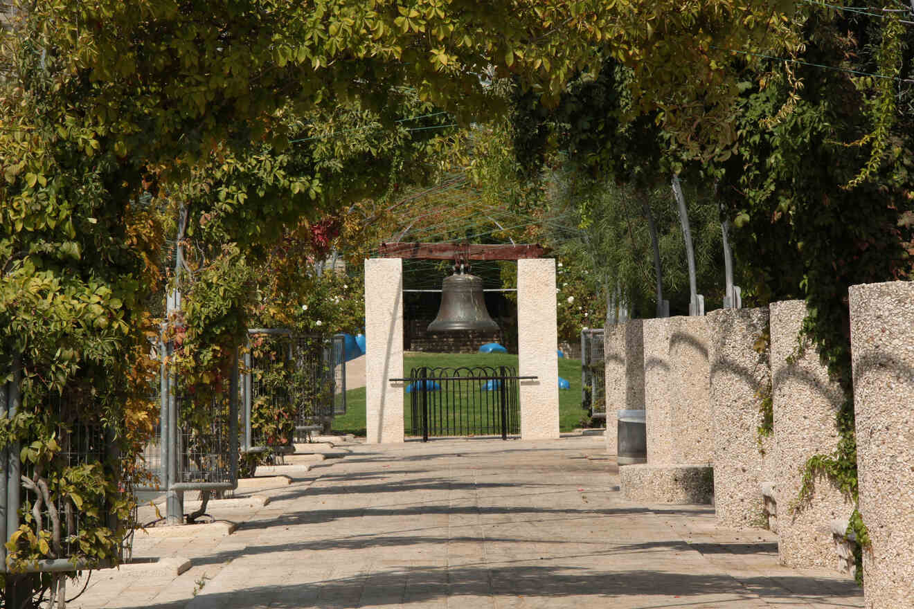 Walkway leading to a large historic bell set within a modern arched structure, surrounded by green foliage.
