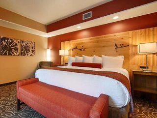 Hotel room with a king-sized bed, decorative wood headboard, two bedside lamps, a red-orange bench at the foot of the bed, and artwork on the wall.