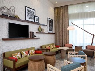 A modern lounge area with green sofas, round wooden tables, a large TV on the wall, and framed art pieces. Decor includes books and vases on shelves. Bright natural light from tall windows.