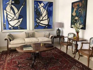 A living room with a beige sofa, two framed abstract paintings, a wooden coffee table, side tables with a lamp and vase of flowers, and two armchairs, all on a red patterned rug.