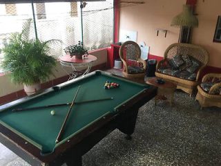 Room with a pool table set up for a game, two wicker chairs with cushions, a small table, a potted plant, and a lamp in the corner.