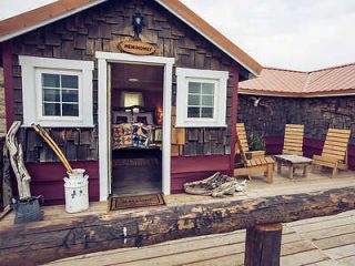 A rustic cabin with a wooden deck, featuring a small porch, two windows, and Adirondack chairs. The cabin door is open, revealing a cozy interior.