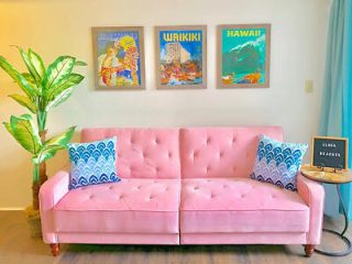 A pink sofa with two blue patterned pillows sits against a wall with three Hawaii-themed posters. A potted plant is on the left, and a small side table with a sign is on the right.