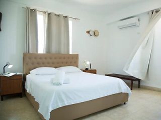 A tidy bedroom with a double bed covered in a white sheet, nightstands, lamps, a bench, air conditioner, and billowing curtains.