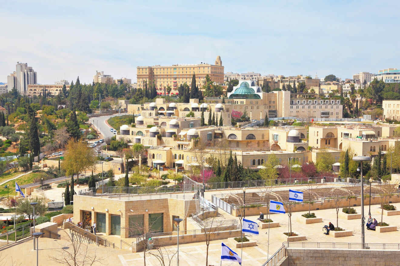 Cityscape showing a mix of modern and traditional architecture with manicured trees and Israeli flags.
