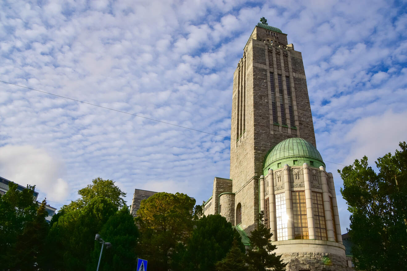 The imposing Kallio Church in Helsinki with its tall, gray stone tower and copper green dome, set against a patterned cloud sky