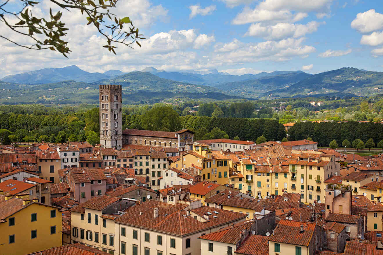 Overlooking view of the ancient town of Lucca, Italy, with its prominent medieval tower rising above the terracotta roofs against the backdrop of lush green hills and blue sky