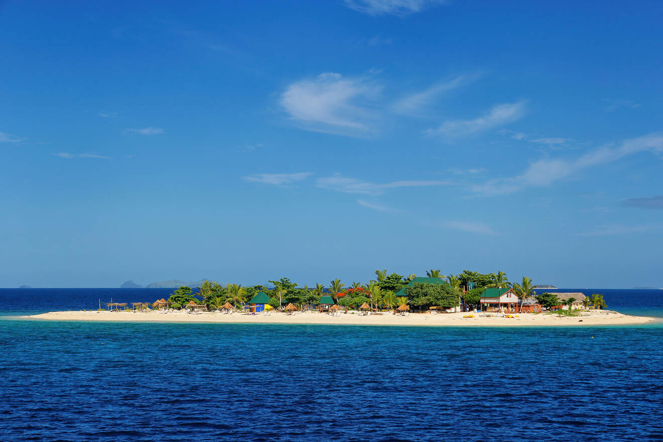 A breathtaking view of a tiny tropical island with a golden sandy beach, palm trees, and a few huts visible, surrounded by the clear blue waters of the ocean.