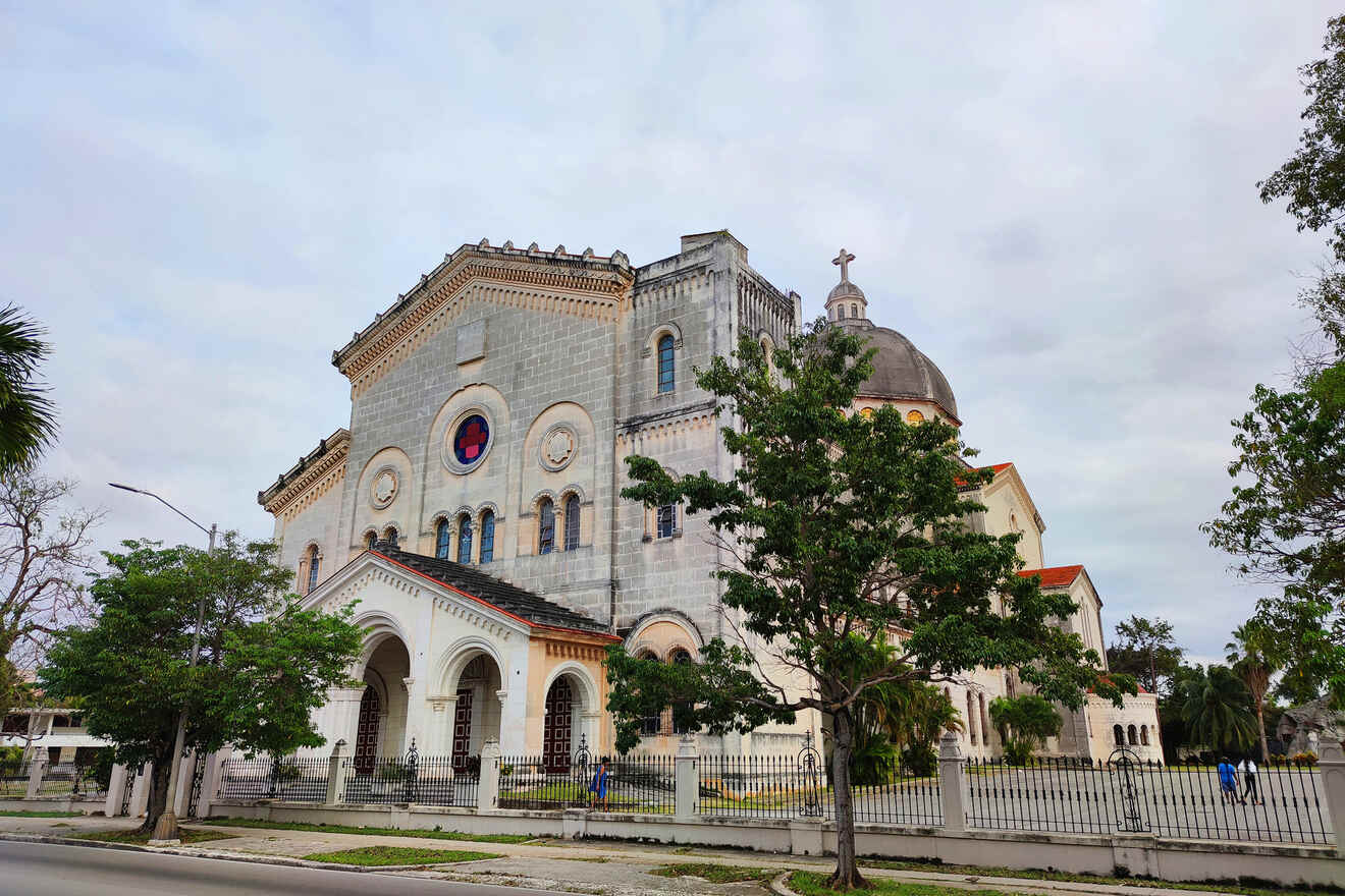 A large stone church with a red rose window and a dome, surrounded by trees and a metal fence, stands on a quiet street under a cloudy sky.