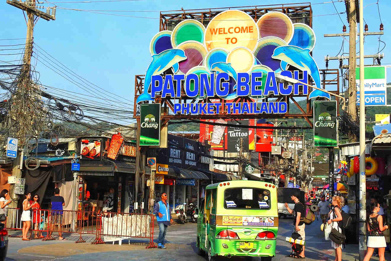 Vibrant street scene at the entrance to Patong Beach in Phuket, Thailand, with colorful signage, bustling tourist activity, and an array of shops and street vendors under a tangle of power lines.

