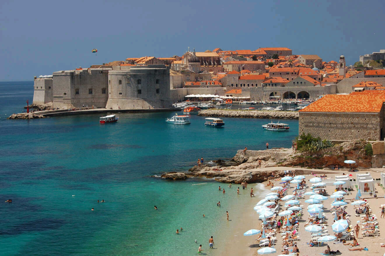 Dubrovnik's bustling Banje Beach with its clear turquoise waters, beachgoers, and the city's historic walls in the background