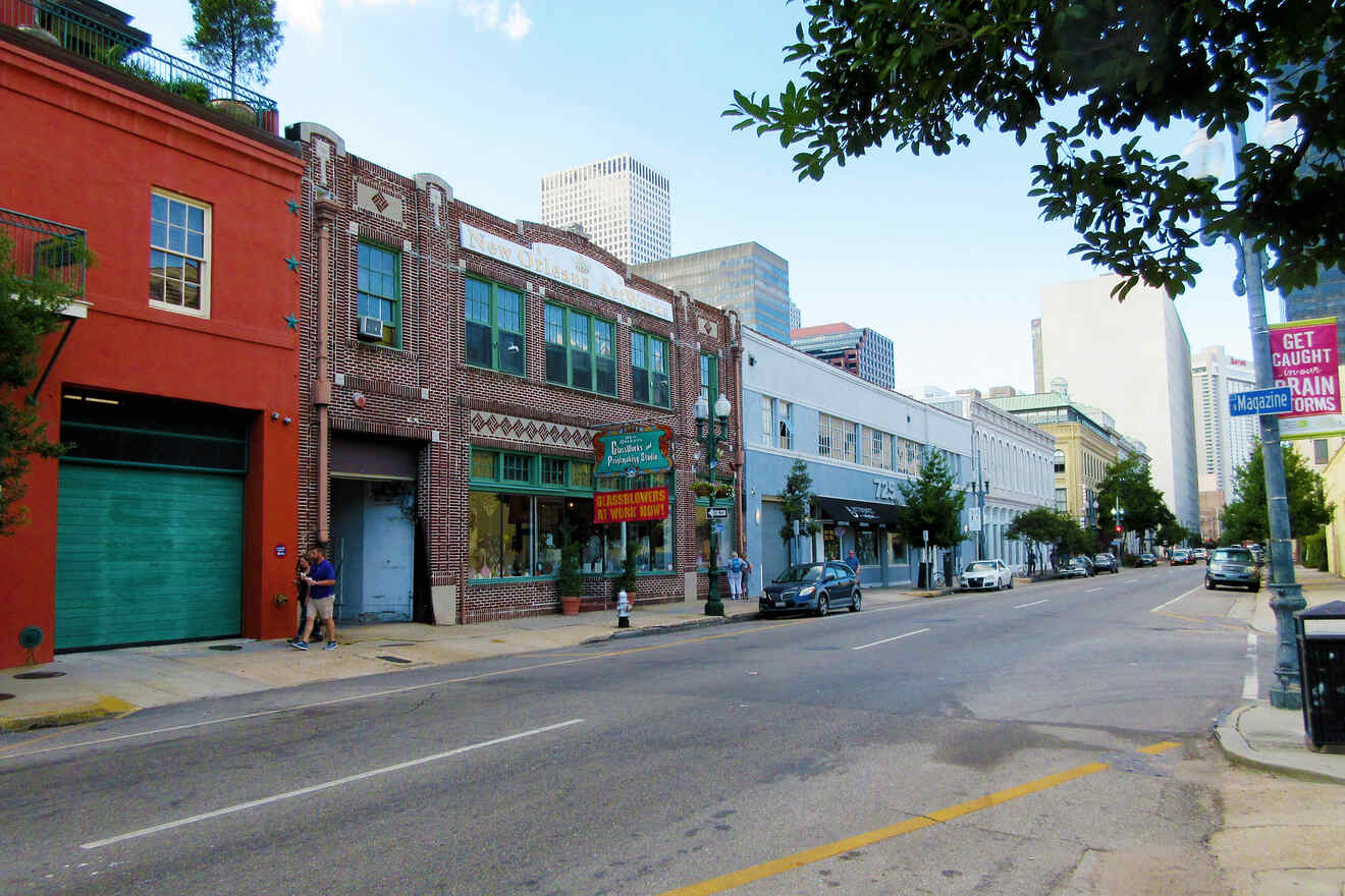 A street in a city lined with buildings, including a red brick establishment with a green sign, cars parked on the side, and a few pedestrians. The skyline is visible in the background.