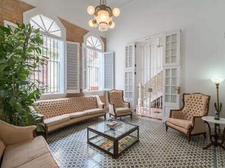 A well-lit room with vintage furniture featuring two sofas, two armchairs, a glass coffee table, and a potted plant. The floor has patterned tiles, and there are windows with shutters and a decorative chandelier.