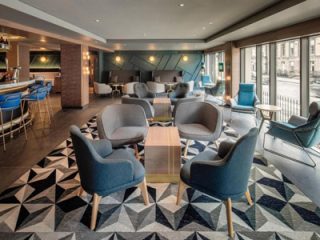 A contemporary hotel lounge with geometric carpeting and plush blue armchairs surrounding sleek wooden coffee tables