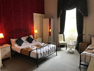 A classic hotel bedroom with two iron-frame beds, red textured wallpaper, bedside lamps, and a view