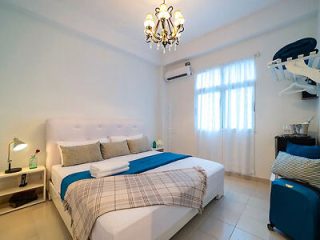 A well-lit bedroom with a double bed, white and blue linens, a bedside lamp, air conditioning unit, suitcase stand, and a chandelier. The room has a window with sheer curtains.