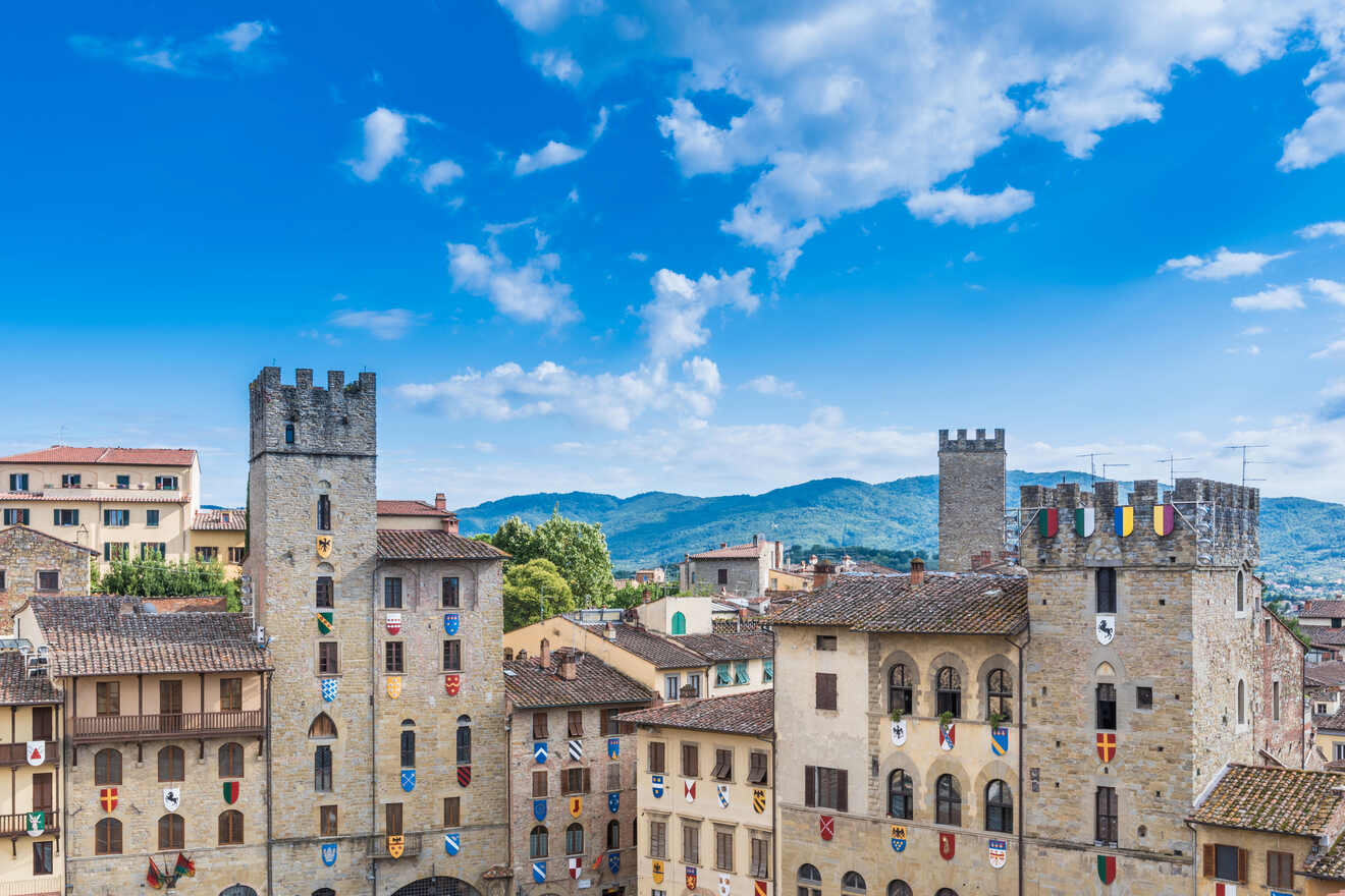 Medieval towers decorated with colorful flags over the stone buildings of Arezzo, Italy, against a backdrop of blue skies and distant mountains