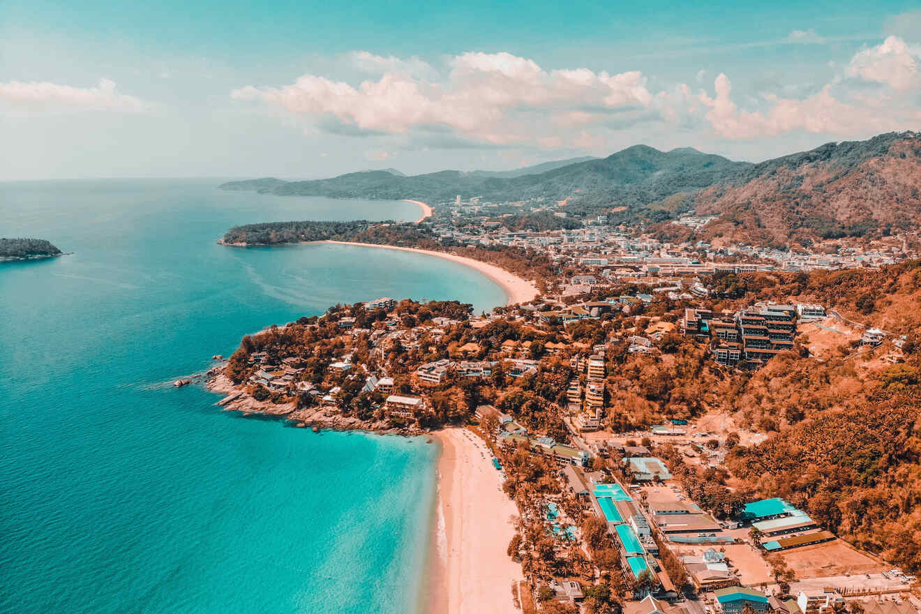 Breathtaking aerial view of a coastal town in Thailand, showcasing a curving sandy beach, aquamarine waters, and a lush mountainous backdrop under a soft blue sky.

