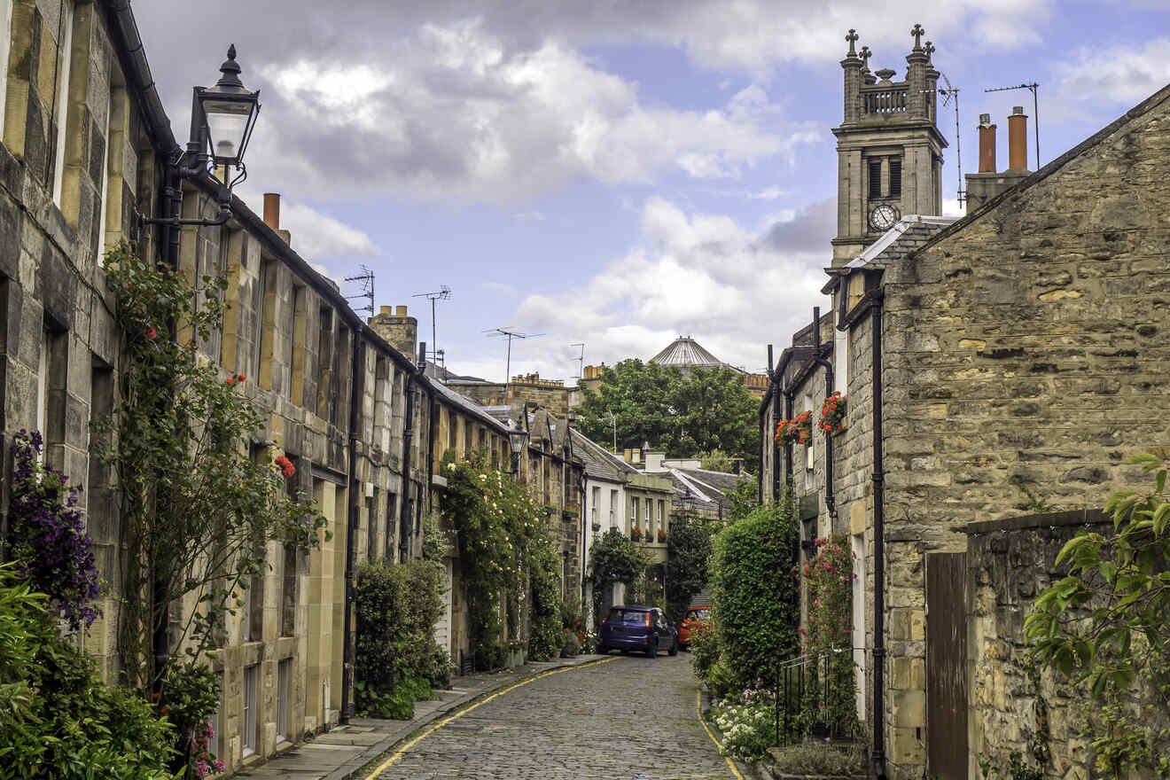 A quaint cobbled lane lined with traditional stone houses adorned with climbing plants and hanging flower baskets, under a sky with fluffy clouds